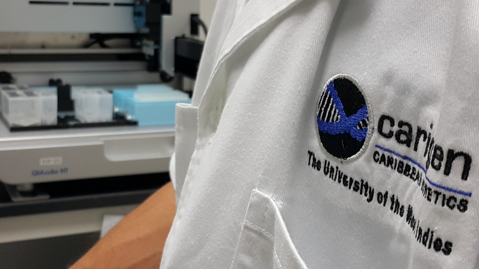 Embroidered CARIGEN Caribbean Genetics logo on a white lab coat, with laboratory automation equipment behind
