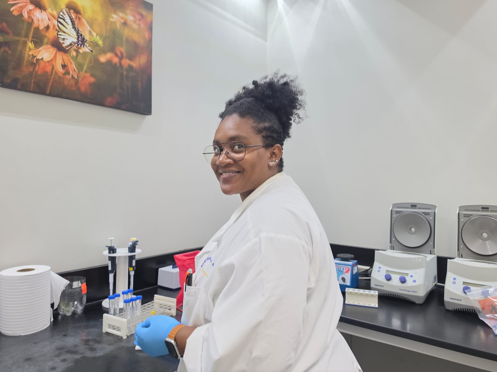 A smiling CARIGEN analyst in a lab coat at the bench with a tube rack, pipettes and benchtop centrifuges behind